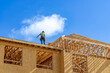 © Andrei Liashchou - A wide horizontal view of a construction worker wearing a safety harness and hard hat standing on a roof deck next to wooden roof trusses during the framing of a multi-story residential building under