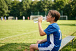 © Meow Creations - Young boy in blue soccer jersey drinking water from clear bottle while resting on sports bench