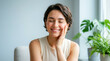 © Iconstarstudio - A young woman with short hair smiles softly, resting her hand on her cheek, seated indoors near a plant.