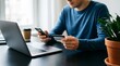 © Mst. Tahamina Akter - Young man using credit card for online shopping payment while holding smartphone and sitting at desk with laptop computer at home during day secure transaction