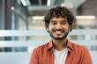 © Liubomir - Young indian man with curly hair and beard smiling confidently in a modern office headshot, conveying friendly professionalism and upbeat startup or corporate energy