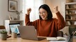 © bayu - A joyful young Asian woman with glasses celebrates a project achievement, raising her fists in triumph while working remotely at her home office desk with a laptop.