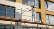 © Pratondo - Professional house painter in white uniform painting the exterior wall of a modern building using a roller on scaffolding.
