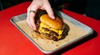© Studio Fiuza - Close-up of a hand picking up a juicy double smash burger from a metal tray on a vibrant red table. High contrast, flash-lit photography perfect for trendy fast food advertising and diner menus.