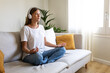 © Daniel - Young woman meditating on the couch listening to guided meditation using wireless headphones. Copy space.