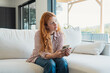 © Daniel - Red-haired woman sitting on a white sofa, holding a smartphone and looking away thoughtfully, relaxing in a bright modern home living room with large windows