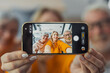 © Daniel - Hand holding a modern smartphone capturing a smiling family selfie with a young woman, senior man, and senior woman creating lasting memories together at home