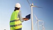© NJF - Female engineer wearing a hard hat and safety vest, looking at wind turbines while holding a laptop, overseeing clean energy production and sustainable development