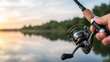 © oliamogdaleva - Close-up of a fisherman holding a spinning rod with a reel over a lake at sunset, focus on fishing gear.
