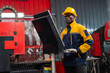 © VStudio - Male worker wears a yellow hard hat and blue-yellow safety jacket. He stands near a black monitor in a factory setting. Red industrial machinery is visible behind him. Focused work environment
