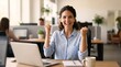 © haemye - Joyful woman celebrates success at her desk. She grips her fists, eyes gleaming with triumph. Laptop rests open before her, notebook beside it. Cafe-style office hums with soft daylight and greenery