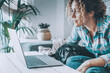© simona - Woman working on laptop at home with her pug on the couch, remote work in a cozy living room with indoor plants