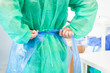 © Alina - A medical worker ties a blue plastic apron over a protective green surgical gown in a clinical setting to ensure safety and hygiene protocols during a medical procedure prep