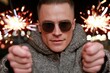 © whitestorm - Man with sunglasses holding sparklers, celebrating with festive lights and bokeh background