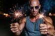 © whitestorm - Muscular bald man with sunglasses holding two sparklers at night with bokeh lights in background