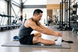 © Meow Creations - Fit asian man stretching his leg on a mat inside a modern gym after a heavy workout