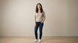 © pro stock photos - Full body shot of a smiling young woman with long wavy brown hair wearing a casual beige sweater dark blue jeans and white sneakers standing confidently with one hand in her pocket against a plain