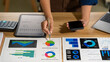 © NanSan - Business professional reviewing charts at a desk, pointing to colorful graphs while holding a phone in a modern office, showing planning, analysis, and financial decision making.