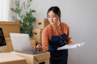 © David - Asian female business owner managing online store operations, checking orders and inventory on laptop while preparing packages for e-commerce shipping