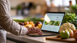 © Tetyana - Person browsing healthy recipes on a laptop, standing in a modern kitchen counter with fresh organic vegetables for cooking and meal preparation