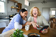© Marko Geber - Caregiver and senior woman laughing over smartphone in home kitchen