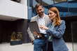 © Novak - Two diverse business professionals standing outside a modern glass office building, looking at a laptop screen and discussing a project during a meeting.
