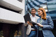 © Novak - Two diverse business professionals standing outside a modern glass office building, looking at a laptop screen and discussing a project during a meeting.