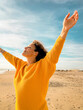© simona - Woman in a yellow sweater standing on a sunny beach with arms raised, eyes closed and smiling, expressing freedom, joy and relaxation by the sea under blue sky and soft sand, healthy lifestyle