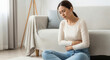 © Iryna - Asian woman sitting on floor holding stomach, suffering from menstrual pain and abdominal discomfort in living room