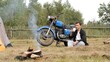 © Aliaksandr - Young man sits by a smoky bonfire drinking coffee beside his tent and parked motorcycle, relaxing on a rural morning during a solo camping trip and countryside adventure