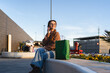 © pablo - Young woman sitting with shopping bags while using a smartphone at an outdoor mall