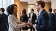 © Artyom - Two professionals exchanging business cards and shaking hands at a networking event with colleagues conversing in the background