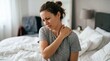 © AlfaSmart - Woman sitting on bed touching her painful shoulder. Girl feeling neck and shoulder ache after sleep. Health problem, muscle strain and body tension concept. Morning at home bedroom.