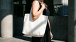 © Studio Fiuza - A woman wearing a black dress carries a blank white canvas tote bag in a sunny urban environment with hard shadows. Perfect for eco-friendly shopping bag mockups, branding.