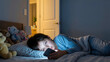 © Przemek - Young boy lying in bed at night using a smartphone in a dark bedroom. The blue light from the screen illuminates his face while he scrolls through his mobile device near teddy bears.