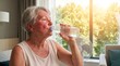 © Olga Ко - Exhausted sweaty senior woman drinking water from a plastic bottle at home in front of a fan during extreme summer heatwave. Elderly dehydration and heat stroke risk concept.