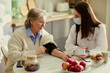 © DragonImages - Senior woman receiving blood pressure check from young female caregiver at home, supporting elder care and home healthcare. Ideal for caregiving, nursing, aging in place marketing