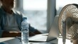 © Evgeniia - Water bottle and desk fan next to laptop in warm office environment. Concept of heatwave preparedness, hydration and maintaining comfort during work