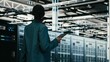© DC Studio - Female programmer walking in data center using tablet to write code, compiling software patches, African American woman in server hub using device application to run programming script, camera B.