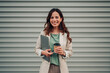 © Zamrznuti tonovi - Young businesswoman smiling, holding a laptop and take away coffee cup, standing against a modern urban background