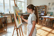 © fanjianhua - A focused teenage female artist in a white apron creates an abstract oil painting on a canvas mounted on an easel inside a bright art studio.