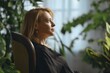 © Aleksei Isachenko/imageBROKER - A woman with long blond hair sits thoughtfully in a wooden chair, surrounded by vibrant green plants in a well-lit room. Sunlight illuminates her face as she gazes into the distance