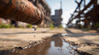 © Mikhail Vorobev - Rusty old metal pipe dripping water onto worn concrete ground with blurred industrial factory structures and bright daylight in the background