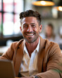 © Kowit - Smiling man in a brown blazer and white shirt sitting indoors, looking confident and approachable with a blurred background featuring warm lighting.