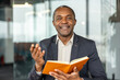 © Liubomir - Mature professional man actively engaging in conversation, holding an open notebook, and smiling confidently in a modern office environment, conveying leadership and successful communication