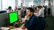 © dotmagic - Man sitting at office desk with green computer screen in modern office setting with coworkers in background