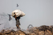 © Travel 'n' Lifestyle - Aminbazar, Bangladesh - 21 February 2026: View of a waste picker carrying a large sack on his head at the Aminbazar landfill with birds flying in the hazy sky.