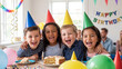 © Bushra - Ecstatic children wearing colorful birthday hats laugh together while posing behind a delicious frosted cake at an indoor party with festive decorations and streamers.