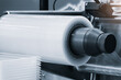 © Aleksandr Matveev - Close-up of a large white roll on industrial machinery. The roll is smooth and glossy, attached to a black mechanical component. Industrial setting features metal parts and precision equipment