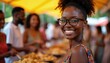 © Pete - Young Black woman wearing glasses smiles at camera, surrounded by people at outdoor food market. She wears a colorful top, standing near serving dishes with prepared meals.